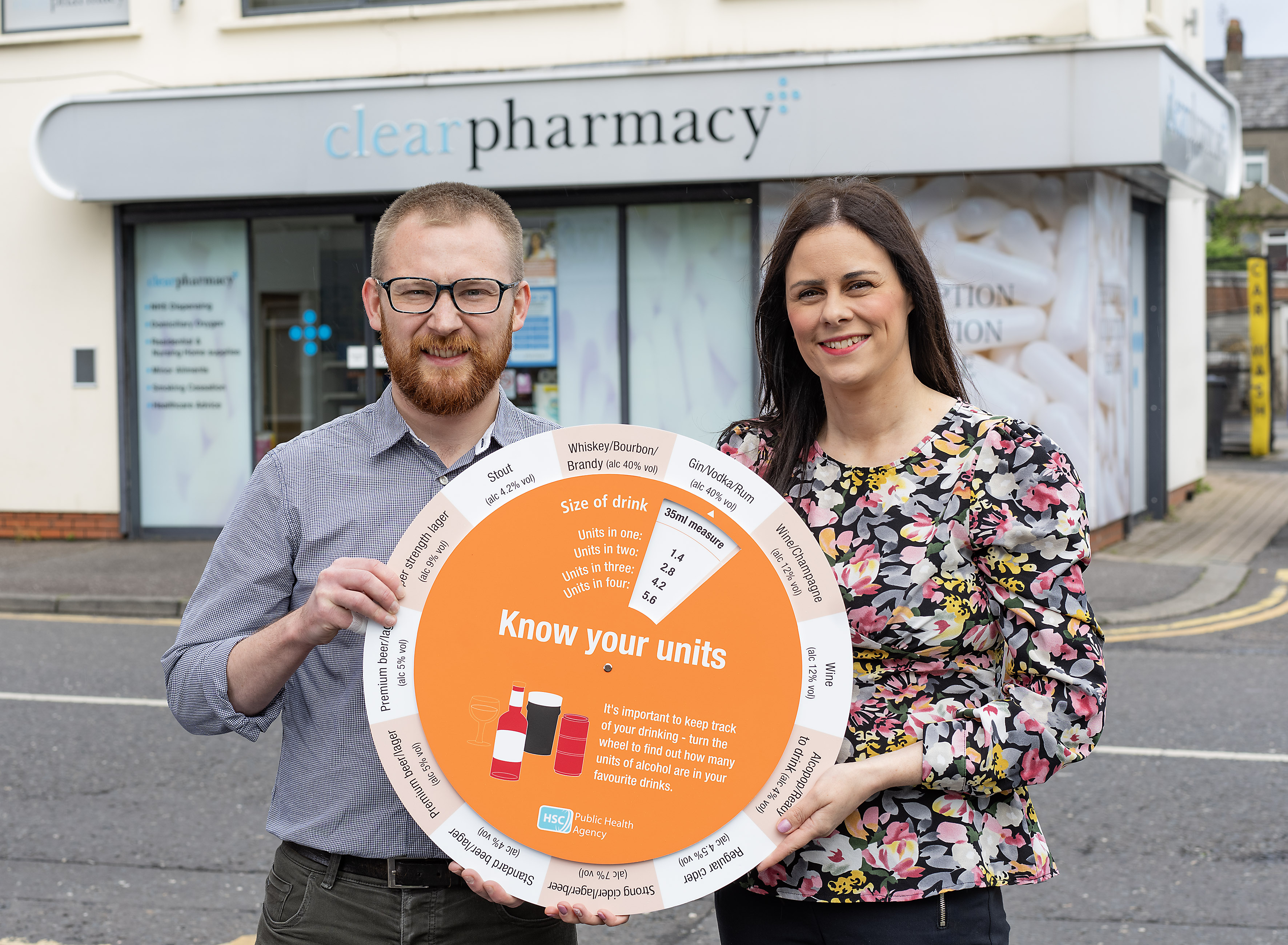 Photograph of PHA rep and pharmacist, holding the 'know your units' campaign unit calculator wheel, outside Clear Pharmacy in Bangor. 